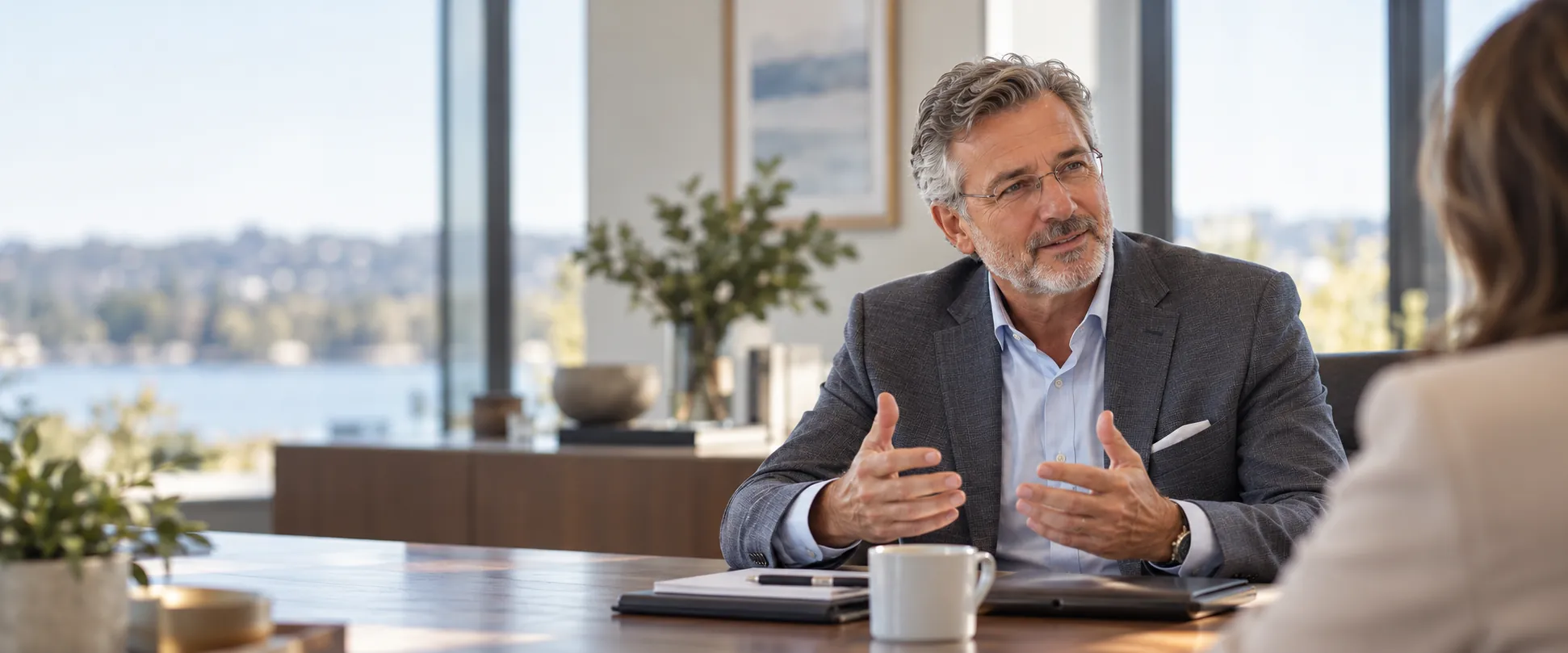 Financial advisor with a grey beard and glasses gesturing with open hands as he talks with a client across his desk