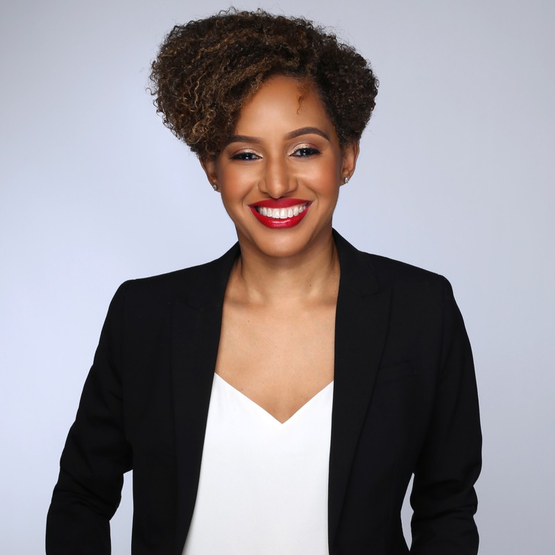 A headshot of a Black woman who has short curly hair. She is wearing a black blazer with a white v-neck shirt underneath