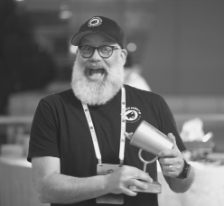 A black and white photo of Matt Halloran from the waist up, holding a trophy. He's wearing a baseball cap and t-shirt. He's grinning widely and has a white beard.